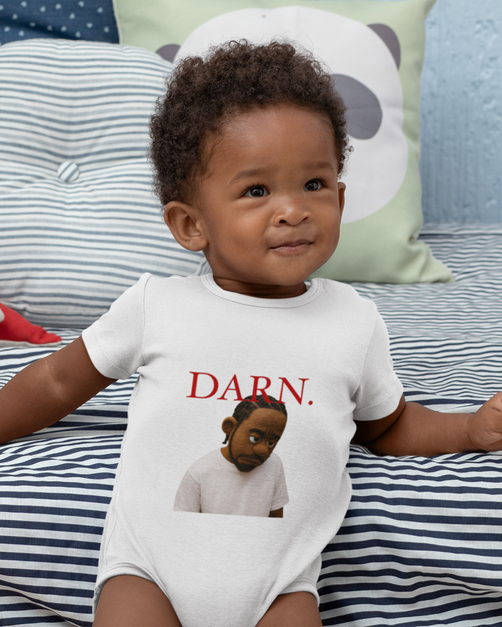 Baby wearing a white onesie with text and an illustration, sitting on a striped bed with toys.