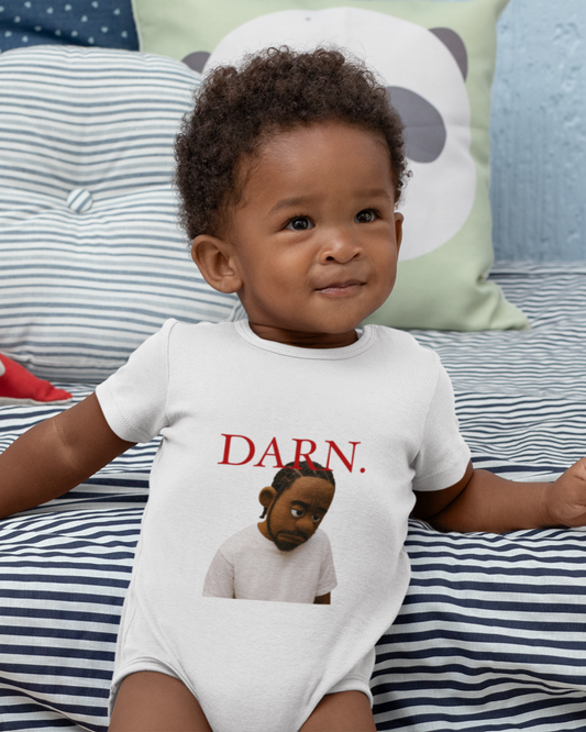 Baby wearing a white onesie with text and an illustration, sitting on a striped bed with toys.